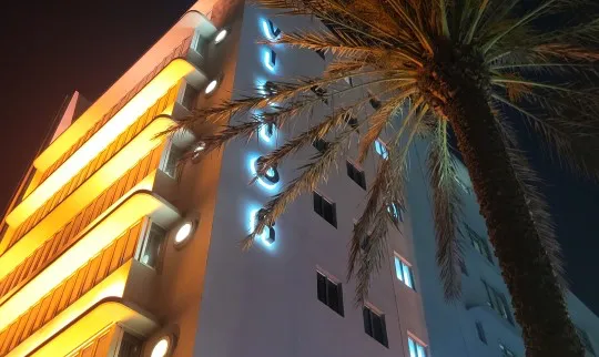 Photo looking up at hi-rise hotel at night with palm tree in foreground, and balcony lighting.